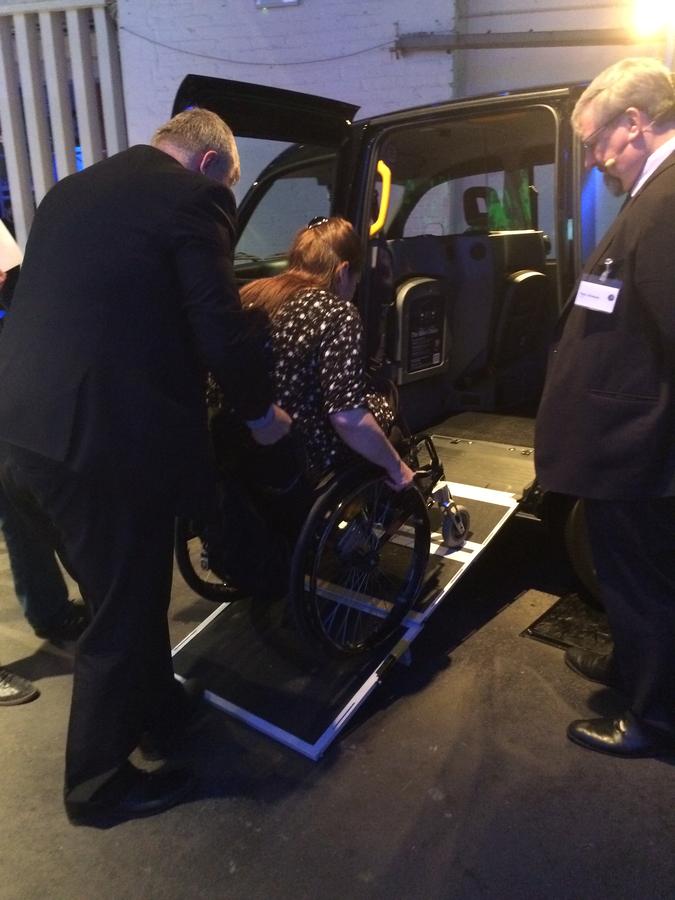 Bild zeigt ein schwarzes London-Taxi von der Seite mit ausgeklappter Rampe. Eine männliche Person schiebt eine Frau im Rollstuhl die Rampe hinauf