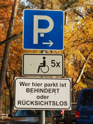 Bild zeigt das Parkschild für den Rollstuhlparkplatz sowie ein weiteres Schild mit der Aufschrift "Wer hier parkt ist behindert oder rücksichtslos"